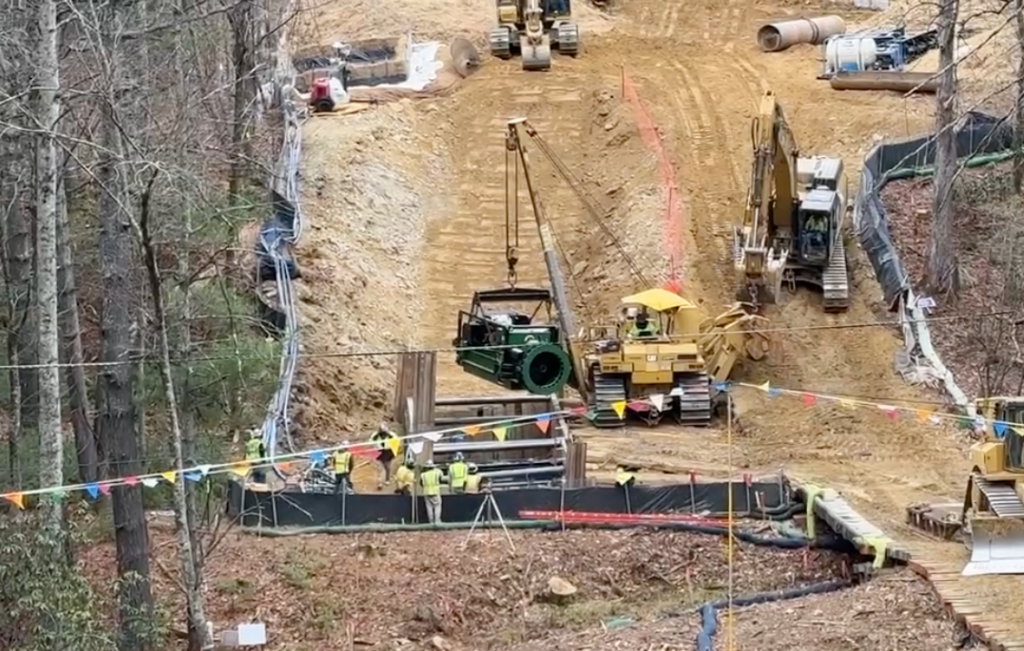 An excavator loads drilling equipment into a bore pit in preparation for tunneling under a creek at the bottom of a steep slope.